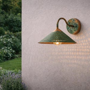 Pode incluir: Luminária de parede para exterior com abajur verde perfurado e braço e placa de parede em bronze. A luz projeta um padrão na parede. Um jardim verde forma o fundo.