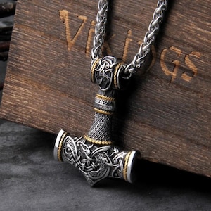 May include: A silver-colored pendant necklace featuring a detailed Thor's hammer design with gold-colored accents. The hammer has intricate patterns and is suspended from a silver chain. The background is a wooden surface with carved text.