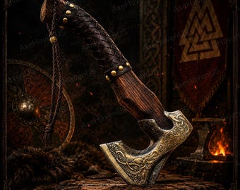 May include: Axe with a wooden handle wrapped in dark brown leather and brass studs. The axe head is gold-colored with intricate carvings. A braided leather cord hangs from the handle. The axe is displayed on a wooden surface.
