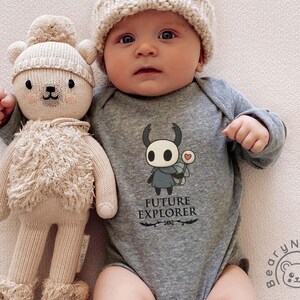 May include: A baby wearing a gray onesie with the text "FUTURE EXPLORER" and a cartoon character. The baby is also wearing a knit cap and holding a beige teddy bear doll.