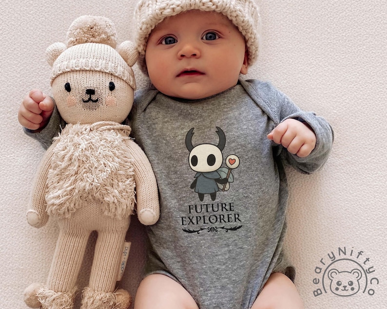 May include: A baby wearing a gray onesie with the text "FUTURE EXPLORER" and a cartoon character. The baby is also wearing a knit cap and holding a beige teddy bear doll.