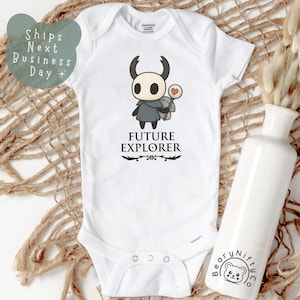 May include: White baby bodysuit with a cartoon character graphic. The graphic features a black and white character with a heart-shaped lollipop and the words "FUTURE EXPLORER" in black. The bodysuit is on a woven surface.