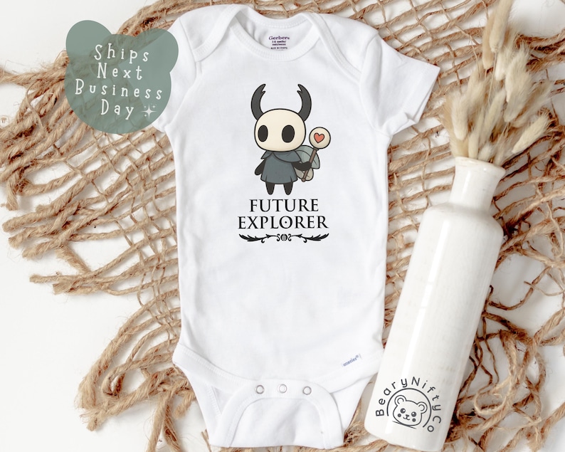 May include: White baby bodysuit with a cartoon character graphic. The graphic features a black and white character with a heart-shaped lollipop and the words "FUTURE EXPLORER" in black. The bodysuit is on a woven surface.