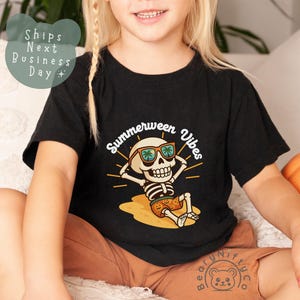 May include: Black t-shirt featuring a cartoon skeleton wearing sunglasses and shorts, with the words "Summerween Vibes." The skeleton is lounging on a beach, with a pumpkin nearby. The shirt is worn by a child.