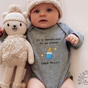 May include: A baby wearing a gray onesie with the text "It's dangerous to go alone. Take this!" and a bottle graphic. The baby is also wearing a cream-colored knit hat and holding a beige teddy bear doll.