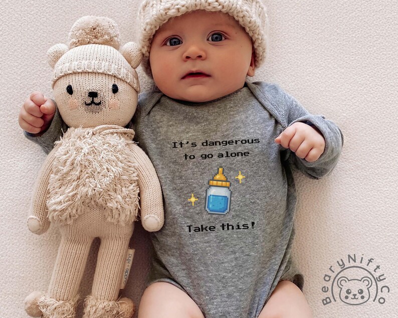 May include: A baby wearing a gray onesie with the text "It's dangerous to go alone. Take this!" and a bottle graphic. The baby is also wearing a cream-colored knit hat and holding a beige teddy bear doll.