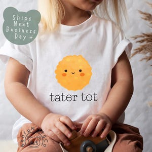 May include: White t-shirt featuring a smiling, cartoon tater tot graphic in shades of orange and yellow. The text "tater tot" is printed below the graphic in black. The shirt is worn by a child.