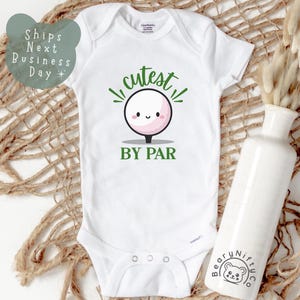 May include: White baby onesie with green text that says "Cutest By Par" and a cartoon golf ball with a pink face.