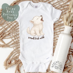 May include: White baby onesie featuring a cartoon wolf cub illustration. The wolf cub is light brown and howling. The text "small but wild" is below the illustration. A white ceramic vase with dried flowers is to the right.