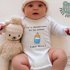 May include: A baby wearing a white onesie with the text "It's dangerous to go alone. Take this!" and a pixelated baby bottle graphic. The baby is also wearing a cream-colored knit hat and is next to a beige teddy bear.