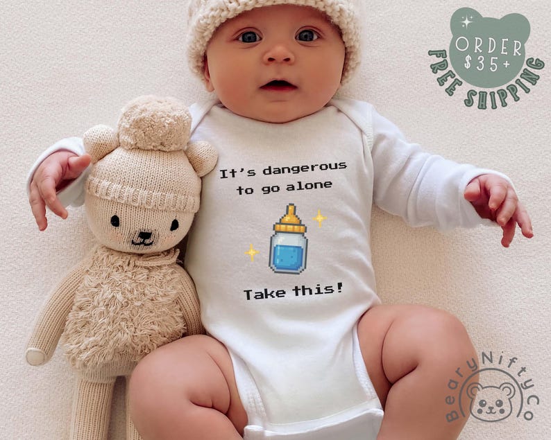 May include: A baby wearing a white onesie with the text "It's dangerous to go alone. Take this!" and a pixelated baby bottle graphic. The baby is also wearing a cream-colored knit hat and is next to a beige teddy bear.