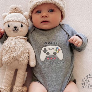 May include: A baby wearing a gray onesie with a video game controller graphic and the text "PLAYER 3 has entered the game." The baby is also wearing a knit cap and is next to a stuffed bear.