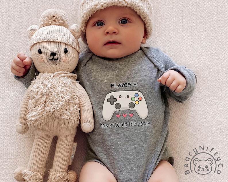 May include: A baby wearing a gray onesie with a video game controller graphic and the text "PLAYER 3 has entered the game." The baby is also wearing a knit cap and is next to a stuffed bear.