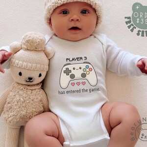 May include: A baby wearing a white onesie with a video game controller graphic and the text "PLAYER 3 has entered the game." The baby is also wearing a knit hat and is lying next to a beige teddy bear.