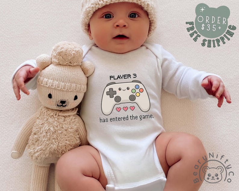 May include: A baby wearing a white onesie with a video game controller graphic and the text "PLAYER 3 has entered the game." The baby is also wearing a knit hat and is lying next to a beige teddy bear.