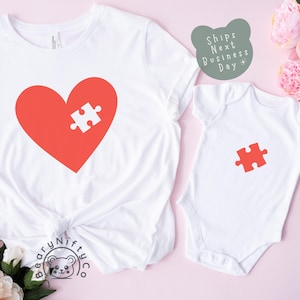 May include: Two white t-shirts with a red heart and puzzle piece design. The larger shirt has a heart with a puzzle piece missing, and the smaller shirt has a single puzzle piece. The shirts are on a pink background.