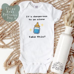 May include: White baby onesie with the text "It's dangerous to go alone. Take this!" and a pixelated baby bottle graphic. The onesie is on a woven surface with a white vase and dried flowers.