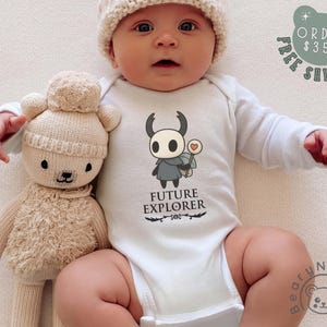 May include: A baby wearing a white onesie with a cartoon character and the words "FUTURE EXPLORER". The baby is also wearing a knit hat and holding a beige teddy bear. The image includes a "FREE SHIPPING" graphic.
