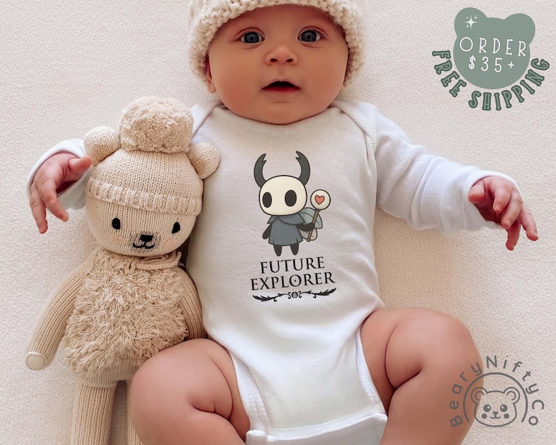 May include: A baby wearing a white onesie with a cartoon character and the words "FUTURE EXPLORER". The baby is also wearing a knit hat and holding a beige teddy bear. The image includes a "FREE SHIPPING" graphic.