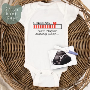 May include: White baby onesie with a red heart and the text "LOADING... New Player Joining Soon..." printed on it. The onesie is lying on a brown wicker basket.