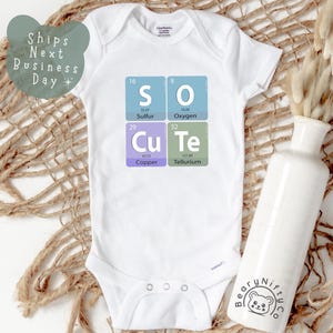 May include: White baby bodysuit featuring the phrase "SO Cu Te" formed from periodic table elements. The garment has a snap closure and short sleeves. A white vase with dried flowers is on the right.