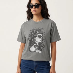 以下が含まれることがあります： サングラスをかけたメデューサの白黒イラストが描かれたグレーのTシャツ。Tシャツはブルージーンズと組み合わせています。モデルも黒いサングラスをかけています。Tシャツはゆったりとしたフィット感です。