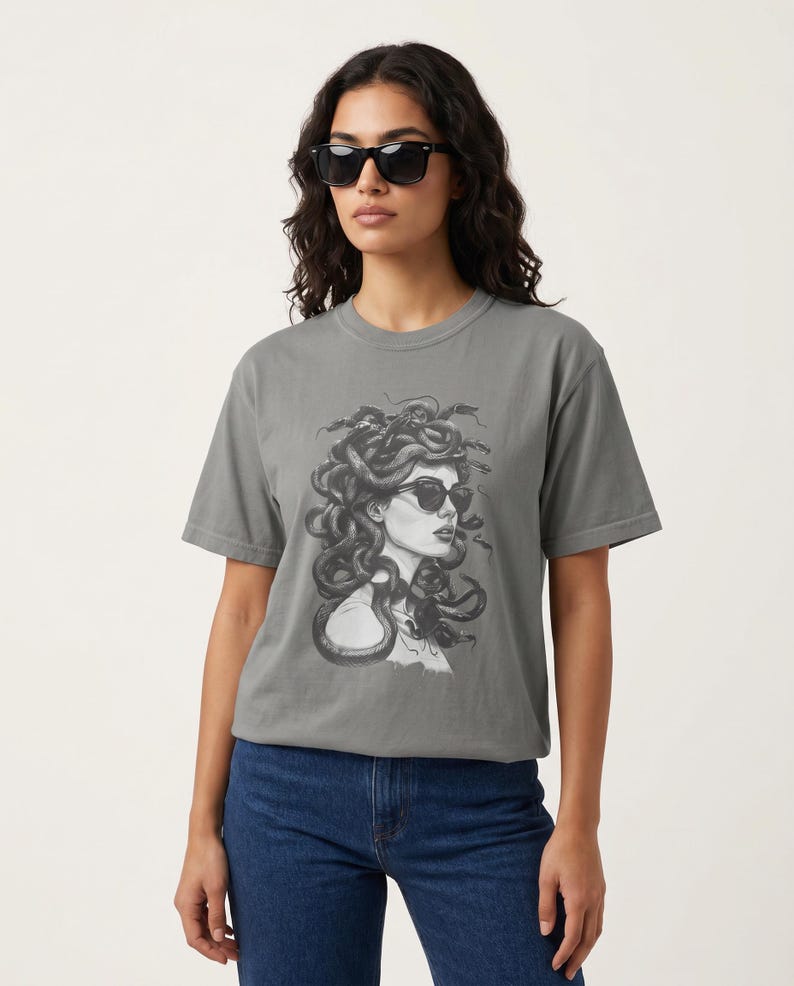 以下が含まれることがあります： サングラスをかけたメデューサの白黒イラストが描かれたグレーのTシャツ。Tシャツはブルージーンズと組み合わせています。モデルも黒いサングラスをかけています。Tシャツはゆったりとしたフィット感です。