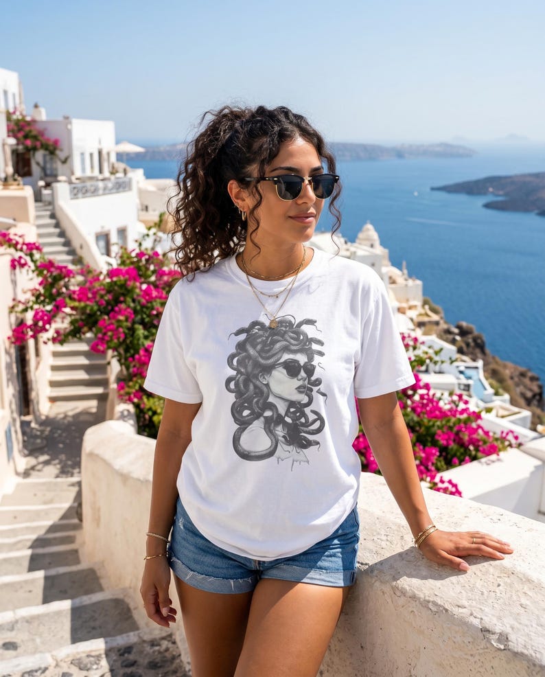 以下が含まれることがあります： サングラスをかけたメデューサの白黒イラストが特徴の白いTシャツ。デニムショーツと合わせています。背景には白い建物、ピンクの花、海の景色があります。