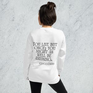 May include: White sweatshirt featuring the quote "YOU LIVE BUT ONCE; YOU MIGHT AS WELL BE AMUSING." in black, attributed to Coco Chanel. The sweatshirt is worn by a person with dark hair and black trousers.