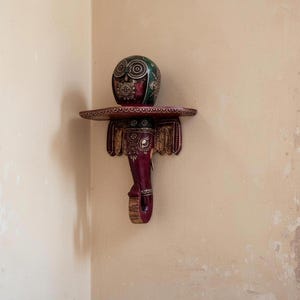 Peut inclure: Étagère murale décorative en forme d'éléphant, de couleur bordeaux avec des détails dorés et des motifs complexes. Un objet décoratif vert et rouge avec un motif circulaire est posé sur l'étagère.