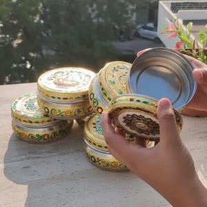 May include: A collection of round metal containers featuring intricate gold-toned filigree patterns with colorful enamel accents. Each container has a removable lid with a central decorative motif. One container is held open to reveal a smooth interior.