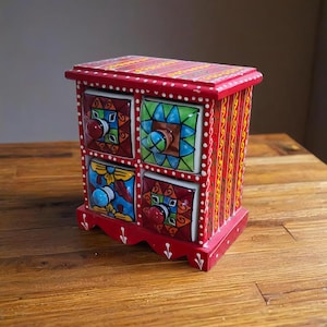 May include: A small, red wooden chest of drawers with four colorful, patterned ceramic drawers. Each drawer has a unique design and a round knob. The chest is decorated with white dots and vertical stripes.