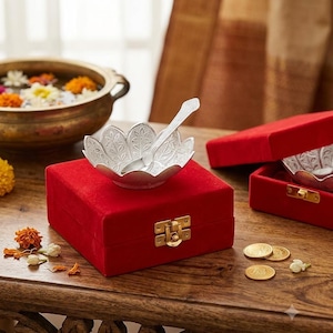 May include: A silver-colored bowl shaped like a lotus flower with an intricate leaf pattern, accompanied by a matching silver-colored spoon. The bowl rests on a square red velvet box featuring a gold-toned metal latch closure on the front side.