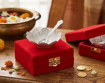May include: A silver-colored bowl shaped like a lotus flower with an intricate leaf pattern, accompanied by a matching silver-colored spoon. The bowl rests on a square red velvet box featuring a gold-toned metal latch closure on the front side.