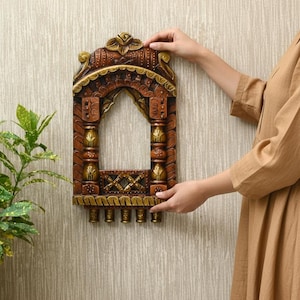 May include: Ornate, wooden wall decor in a window frame design. The piece features intricate carvings, with a mix of brown and gold tones. The decorative item is held against a textured, neutral-toned wall.