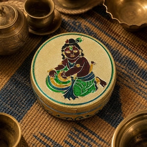 May include: A round gold-toned metal box featuring an intricate enamel illustration of a dark-skinned figure with a peacock feather in their hair, wearing green and blue garments, while reaching into a golden bowl with a textured background.