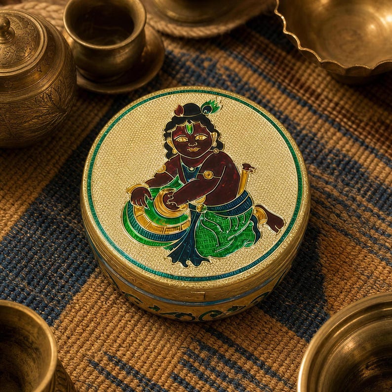 May include: A round gold-toned metal box featuring an intricate enamel illustration of a dark-skinned figure with a peacock feather in their hair, wearing green and blue garments, while reaching into a golden bowl with a textured background.