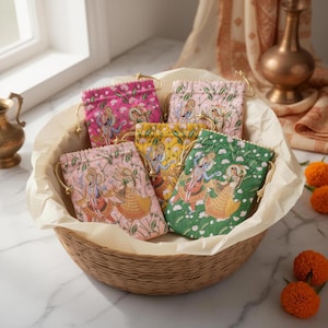 May include: A wicker basket contains five patterned drawstring bags. The bags, in pink, yellow, and green, depict figures in traditional dress. Each bag has a gold-coloured drawstring. The basket sits on a marble surface.