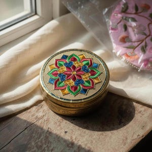May include: A round brass container featuring a textured gold-tone surface and a central lid design with a colorful floral mandala pattern in shades of red, green, blue, and yellow. The sides of the container are engraved with a repeating scroll motif.