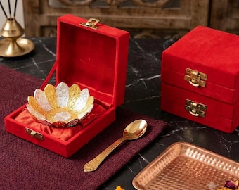 May include: A decorative bowl with a lotus-like petal design in alternating silver and gold tones, presented inside a red velvet box with a gold-tone metal latch. A matching gold-tone spoon and a rectangular hammered copper tray are also included.