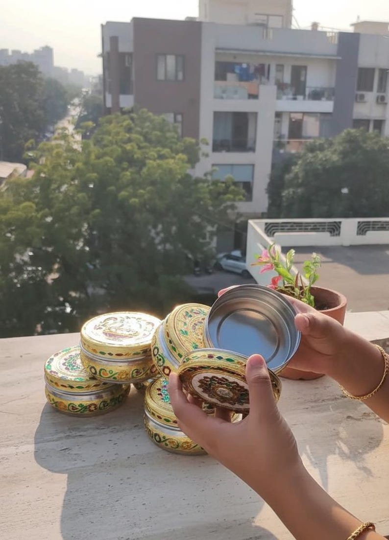 May include: A collection of round metal containers featuring intricate gold-toned filigree patterns with colorful enamel accents. Each container has a removable lid with a central decorative motif. One container is held open to reveal a smooth interior.