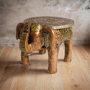 May include: A decorative wooden elephant stool with a round top. The stool is painted in shades of gold, copper, and green, with intricate carved details. The elephant's head forms the front of the stool, and its legs support the seat. The stool measures approximately 12 inches in height.