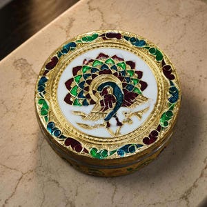 May include: A round gold-tone metal trinket box featuring a central enamel inlay of a peacock with a fanned tail in shades of green, blue, and deep red against a white background. The lid border has intricate gold filigree and colorful enamel.