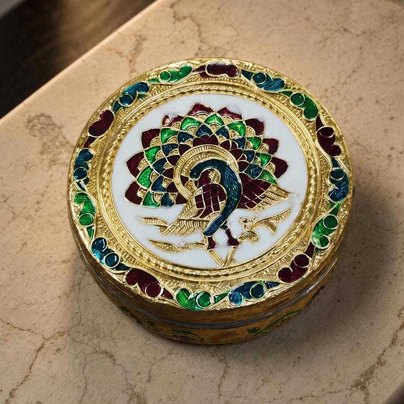 May include: A round gold-tone metal trinket box featuring a central enamel inlay of a peacock with a fanned tail in shades of green, blue, and deep red against a white background. The lid border has intricate gold filigree and colorful enamel.