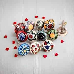 May include: A selection of ceramic cabinet knobs with floral patterns in shades of red, blue, yellow, and green. The knobs are displayed on a light grey surface, accompanied by small red heart decorations.