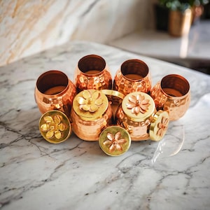 May include: Copper-coloured metal jars with a hammered texture and flower-shaped lids. The jars are arranged on a marble surface. Some lids are off, revealing the interior. The lids and jars have a warm, metallic finish.