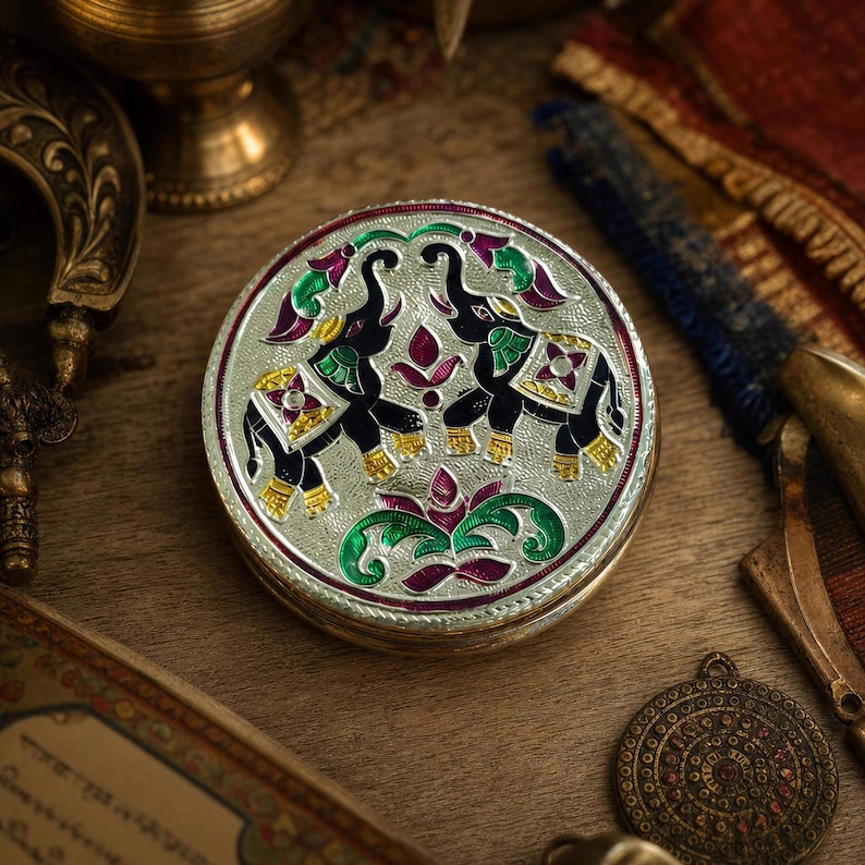 May include: A round silver-toned metal box featuring an intricate embossed design of two black elephants facing each other. The elephants are decorated with vibrant green, purple, and yellow enamel accents, surrounded by floral motifs and patterns.