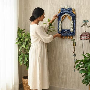 May include: A decorative, blue and gold wooden wall hanging with an arched window design. The wall hanging has intricate carvings and is being placed on a textured wall. A woman in a cream-colored dress is adjusting the wall hanging.