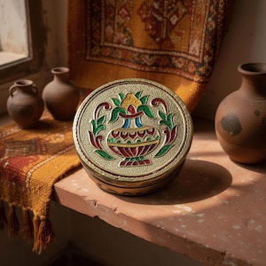 May include: A round brass container with a lid featuring an embossed and enameled design of a kalash pot. The central motif is colored in red, green, yellow, and blue, set against a textured gold-toned metal surface with intricate patterns.