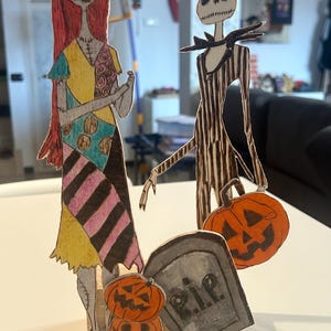 Può includere: Sagome di cartone di Sally e Jack Skellington da The Nightmare Before Christmas, con una lapide e zucche. Sally ha i capelli rossi e un vestito patchwork. Jack indossa un abito gessato. Le zucche sono arancioni.
